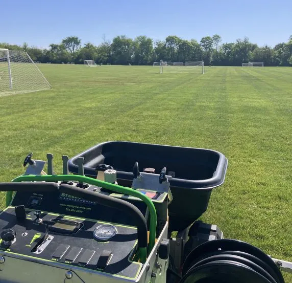 A Grant Keys Lawn Services commercial fertilizer spreader applies treatment to a well-maintained soccer field, with multiple goals visible across the lush green turf under a clear blue sky.
